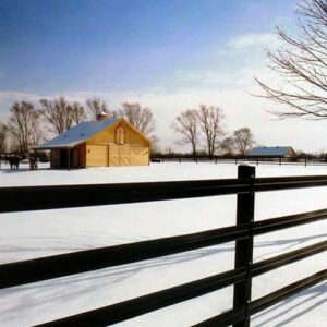 brown fence in a snow field with a tan barn in the background