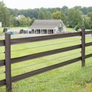 3 rails of brown 525 flex fence in a green field. There is a stable in the background and the fence rails have wire fence between each rail.