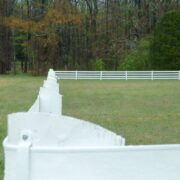 White flex fence in a green filed that borders a forest