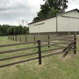 a brown 4 rail fence in front of a large tan barn