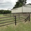 a brown 4 rail fence in front of a large tan barn
