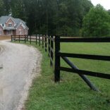 a three rail fence bordering a curving road with a luxury brick home in the background