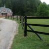 a three rail fence bordering a curving road with a luxury brick home in the background